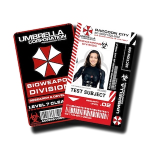 Könnte beinhalten: Zwei Ausweise mit dem Umbrella Corporation Logo. Eine Karte ist schwarz und rot, mit dem Text "Bio Weapons Division" und "Level 7". Die andere Karte ist rot und weiß, mit einem Foto einer Frau und dem Text "Test Subject".