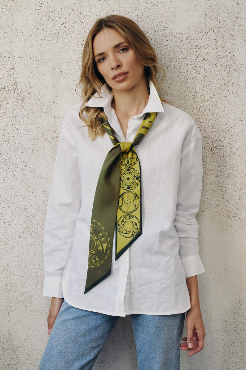 May include: A woman wearing a white linen shirt with a green and gold patterned scarf tied around the collar. The scarf has a geometric design.