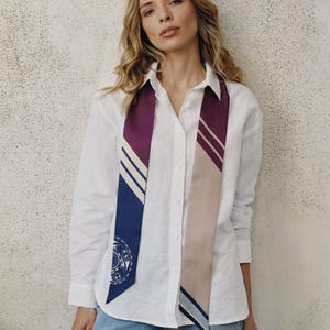 May include: A woman wearing a white button-down shirt and blue jeans. A patterned scarf in shades of purple, blue, and beige hangs from the neck. The scarf features geometric designs and stripes.