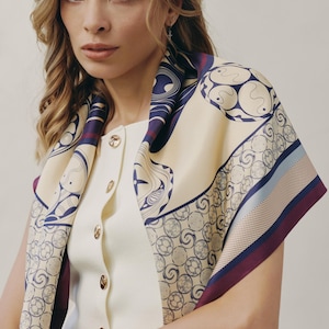May include: Cream-colored silk scarf with a navy blue and burgundy geometric pattern. The scarf is draped over a white top with gold buttons. The scarf's design features circular and curved shapes.