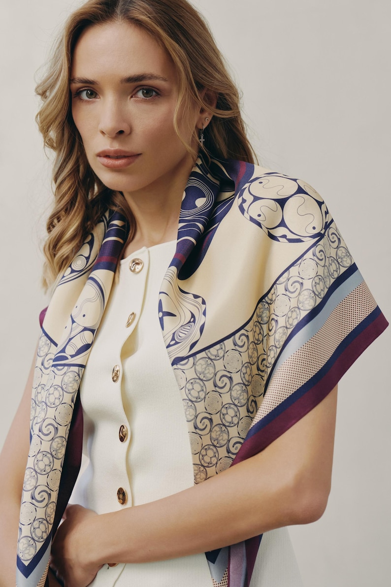May include: Cream-colored silk scarf with a navy blue and burgundy geometric pattern. The scarf is draped over a white top with gold buttons. The scarf's design features circular and curved shapes.