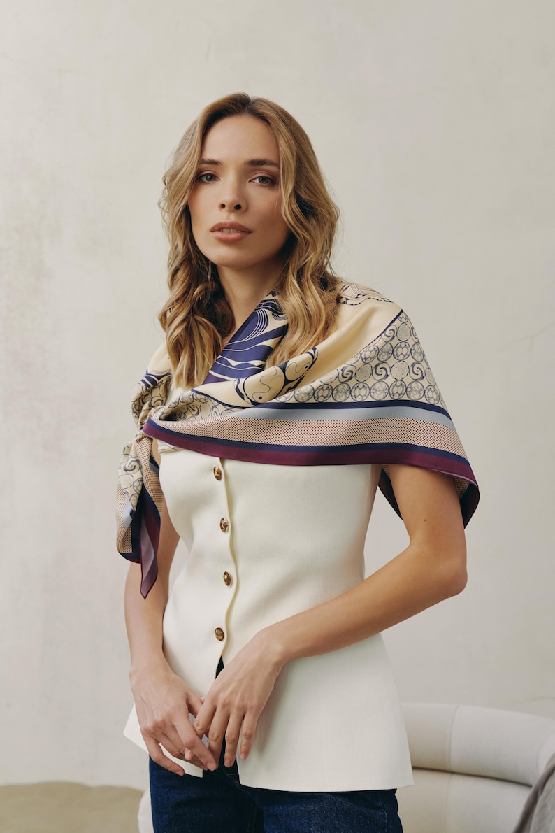 May include: A woman wearing a cream-colored button-down vest and a patterned silk scarf. The scarf features a navy blue and beige design with a burgundy border. The vest has gold-colored buttons.