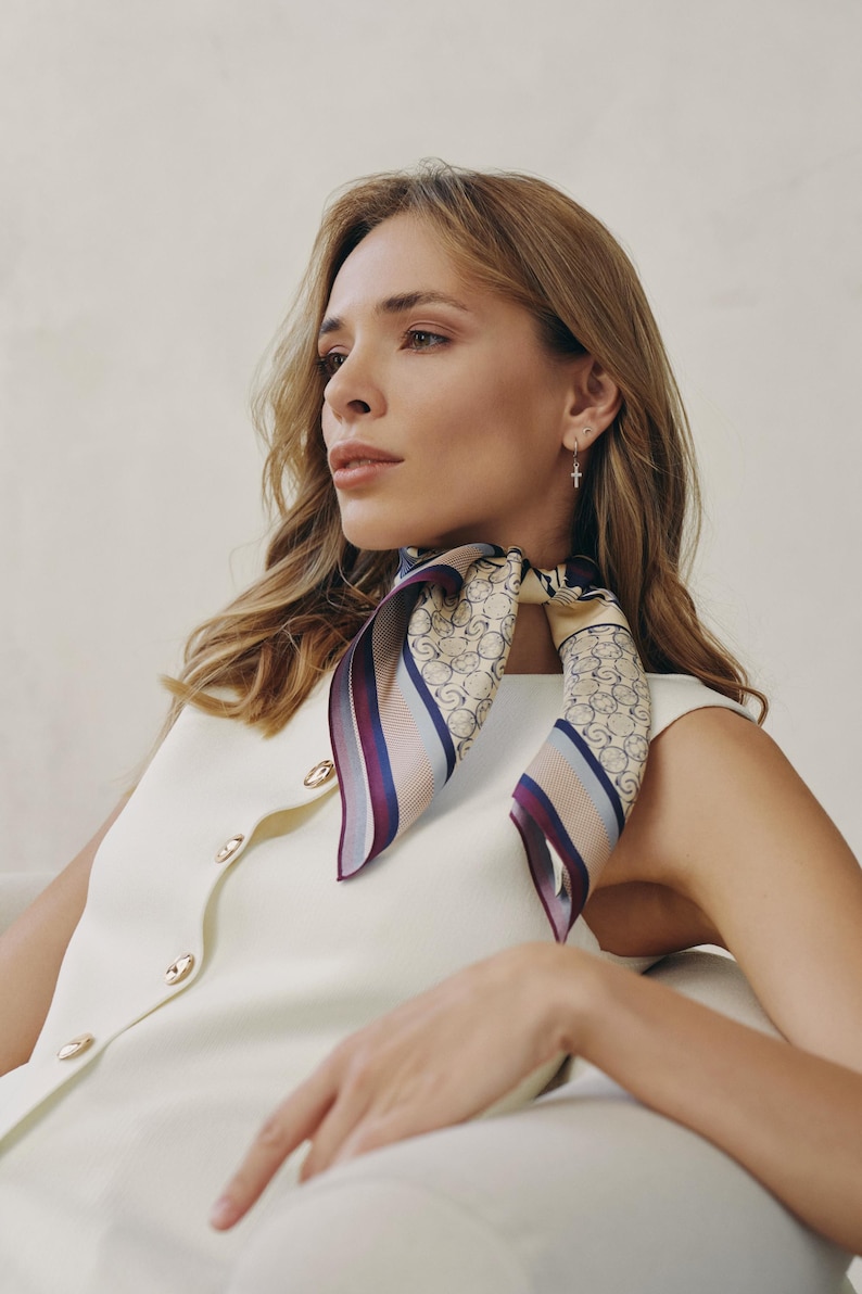 May include: A woman wearing a cream-colored dress and a patterned silk scarf. The scarf features a beige background with a repeating circular design, accented by stripes of burgundy and blue. Gold earrings are visible.