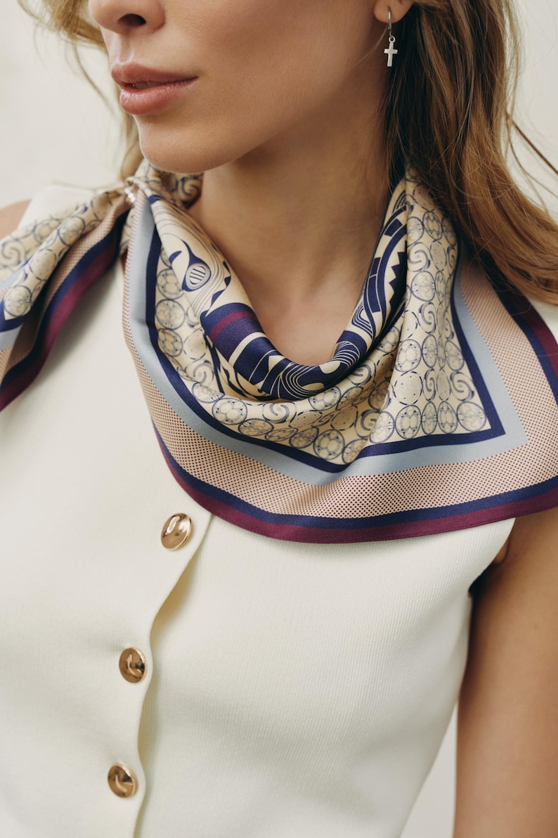 May include: A silk scarf with a geometric pattern in beige, navy, and burgundy is draped around the neck. The scarf is paired with a cream-colored sleeveless top with gold buttons. The scarf's design includes circles and other shapes.
