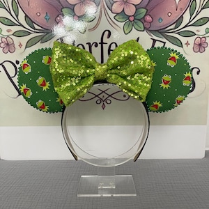 Puede incluir: Orejas de ratón verdes y amarillas con un lazo de lentejuelas verdes. Las orejas presentan un personaje de rana de dibujos animados.