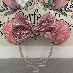 May include: Pink sequined bow and ears headband. The ears are pink with a repeating cartoon character pattern. The headband is clear plastic. The bow is made of pink sequins.