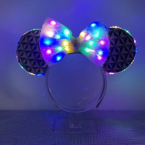 Puede incluir: Diadema blanca con un gran lazo blanco y dos orejas blancas con un patrón geométrico. El lazo y las orejas tienen luces LED multicolores integradas.