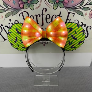 May include: A black headband with attached ears and a bow. The ears are green with yellow stripes and purple bat silhouettes. The bow is orange and yellow plaid with small lights. The text "Perfect Ears" is visible in the background.