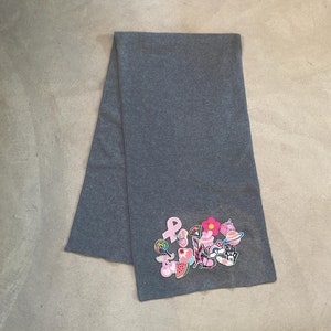 Può includere: Una sciarpa grigia con una varietà di toppe colorate cucite, tra cui un nastro rosa, un'anguria, un fiore, una farfalla e un cupcake.