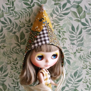 May include: A doll with blue eyes and long, light brown hair wearing a patchwork hat with a yellow, brown, and floral pattern. The doll is wearing a yellow dress and has a cream-colored scarf. The background is a green and white leaf pattern.