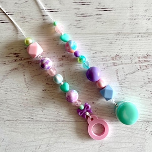 May include: Two beaded pacifier clips with pastel-colored beads. One clip features pink, purple, and teal beads, a pink bow, and a pink ring. The other clip has pink, purple, teal, and blue beads.