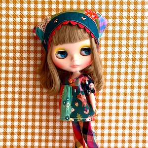 May include: A doll with shoulder-length brown hair and blue eyes wearing a teal headscarf with a scalloped edge, a dark green tunic featuring a colorful woodland animal print, and plaid trousers in shades of red, yellow, and blue with red platform shoes.