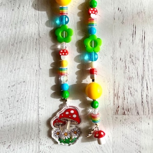May include: Two beaded hanging ornaments. Each ornament features a yellow sphere, green flower-shaped beads, and colorful beads. One ornament has a mushroom charm and a mushroom and daisy charm.