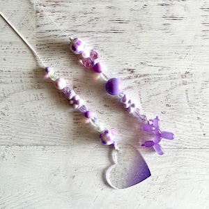 May include: Two beaded wands with purple and white beads, a heart-shaped charm with a gradient effect, and a purple balloon animal. The wands are on a white wooden surface.