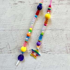 May include: Two colorful beaded phone charms with rainbow accents. One charm spells "BLYTHE" and has a lollipop at the end. The other charm spells "DOLLY" and has a rainbow star at the end.