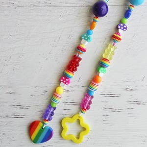 May include: Two colorful beaded accessories with rainbow, heart, and star charms. The beads are in various shapes and colors, including purple, yellow, red, and blue. The accessories have a playful and whimsical design.
