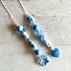 May include: Two beaded accessories with a light blue and white color scheme. Each accessory features a variety of beads, including round beads, butterfly-shaped beads, and heart-shaped beads. One accessory has a star-shaped charm. The accessories are strung on white cords.
