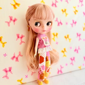 May include: A doll with long, light pink hair and bangs, wearing a pink top with a bunny graphic and patchwork pants in pink, orange, and yellow. The doll has yellow shoes and is set against a backdrop with balloon animal patterns.