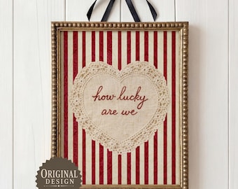 May include: A rectangular framed print featuring a red and white vertical striped background with a central cream-colored heart-shaped lace doily. The text "how lucky are we" is embroidered in red script in the center of the heart. A black ribbon bow is attached to the top of the frame.