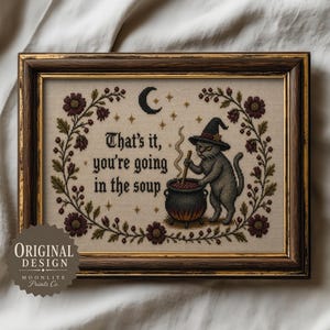 Puede incluir: Una pieza de punto de cruz enmarcada que muestra un gato gris con sombrero de bruja revolviendo un caldero burbujeante. El texto dice, That's it, you're going in the soup. El diseño está rodeado por una corona floral con luna y estrellas.