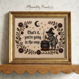 May include: A framed cross-stitch embroidery featuring a grey cat wearing a witch hat, stirring a black cauldron. The text reads, "That's it, you're going in the soup." The design is surrounded by a floral wreath, stars, and a crescent moon.