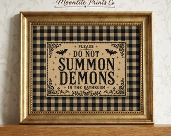 May include: A framed wall art piece featuring a tan rectangular sign with black text that reads PLEASE DO NOT SUMMON DEMONS IN THE BATHROOM. The sign includes black bat and skull illustrations, set against a black and tan buffalo check background.