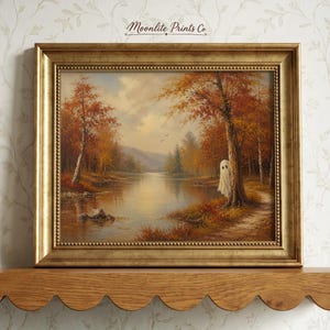 May include: A framed landscape painting in a gold frame featuring an autumn forest scene with a lake and a small white ghost figure standing by a tree. The text Moonlite Prints Co is printed at the top center of the artwork in a dark script font.