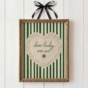 May include: A framed picture with a green and white striped background. A heart-shaped doily features the text "how lucky are we" and a shamrock. The frame is gold-coloured, and a black ribbon is tied at the top.