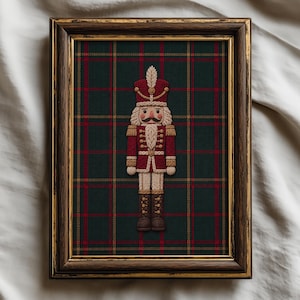 May include: A framed embroidery piece featuring a nutcracker figure centred on a dark green and red plaid fabric background. The nutcracker wears a red uniform with gold accents, a tall hat with a feather, and brown boots, all encased in a dark wood frame.