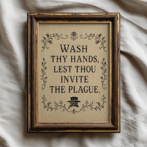 May include: A framed cross-stitch embroidery featuring the black text WASH THY HANDS, LEST THOU INVITE THE PLAGUE. centred on a tan fabric background. The design includes a black plague doctor mask icon and decorative floral borders in a dark wood frame.