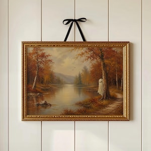 May include: A landscape painting in a gold ornate frame hanging by a black ribbon. The artwork depicts an autumn forest scene with a calm lake, orange and brown trees, and a small white ghost figure standing behind a tree trunk near the water.