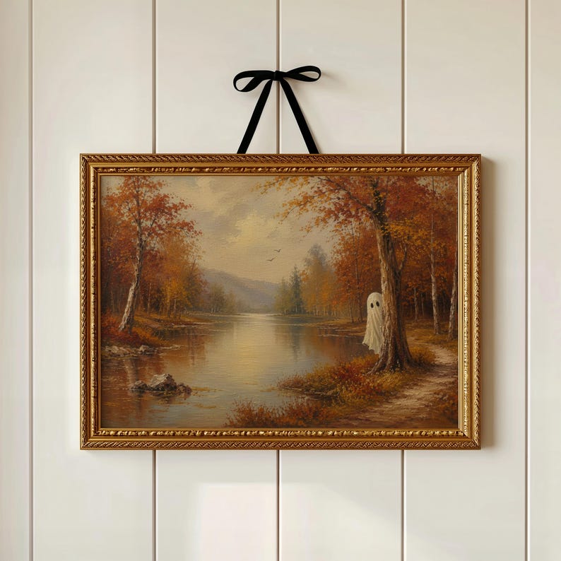 May include: A landscape painting in a gold ornate frame hanging by a black ribbon. The artwork depicts an autumn forest scene with a calm lake, orange and brown trees, and a small white ghost figure standing behind a tree trunk near the water.