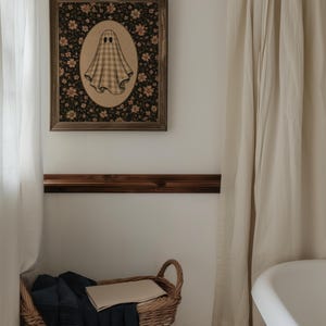 Może przedstawiać: Oprawiona prostokątna dekoracja ścienna z centralną owalną ilustracją ducha w beżową kratkę. Duch znajduje się na ciemnym tle wypełnionym drobnymi r&oacute;żowymi i białymi motywami kwiatowymi, zamknięty w drewnianej ramie.