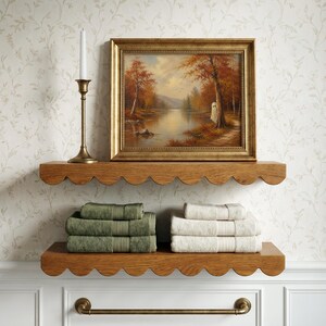 May include: Two scalloped oak shelves mounted on a wall, holding a framed landscape painting of an autumn forest with a ghost, a brass candlestick, and two stacks of folded towels in olive green and cream, with a brass towel bar underneath.