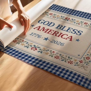 May include: A patriotic design featuring the text "GOD BLESS AMERICA" with "1776-2026" and star accents. The design is framed with floral and star patterns, and a blue gingham border. The image shows a person unrolling the design.