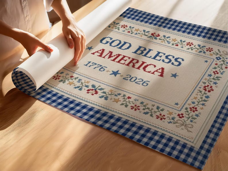 May include: A patriotic design featuring the text "GOD BLESS AMERICA" with "1776-2026" and star accents. The design is framed with floral and star patterns, and a blue gingham border. The image shows a person unrolling the design.