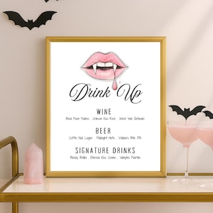 May include: A framed sign with a gold border features a pink lip design with fangs and the words "Drink Up." Below, it lists wine, beer, and signature drinks. Black bat cutouts decorate the top. The sign is on a gold shelf with a pink crystal.