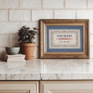 May include: A framed artwork with a gold-colored frame. The artwork features the text "GOD BLESS AMERICA" with the years "1776-2026" and a floral border. The background is a blue and white gingham pattern.