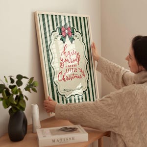 May include: A framed vertical wall art print featuring a dark green and white vertical striped background. The center displays a decorative white scalloped frame with a red bow at the top and the red text have yourself a merry little Christmas.