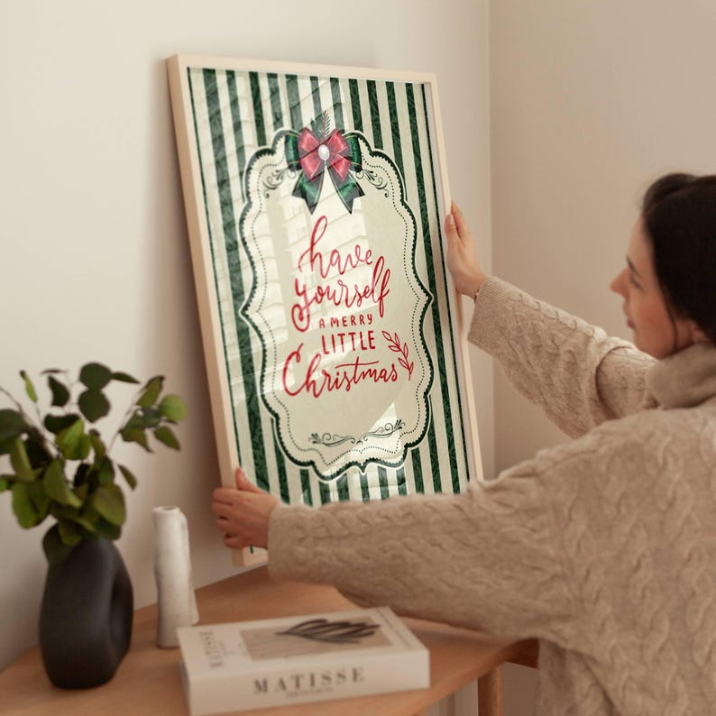 May include: A framed vertical wall art print featuring a dark green and white vertical striped background. The center displays a decorative white scalloped frame with a red bow at the top and the red text have yourself a merry little Christmas.