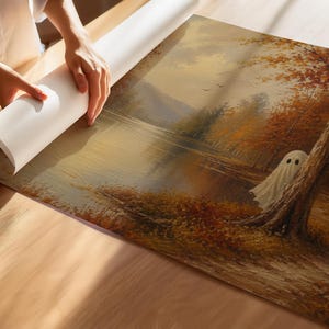 May include: A rectangular canvas print featuring an oil painting of an autumn landscape with a lake, orange and brown trees, and a small white ghost figure standing near a tree trunk. The artwork is being unrolled on a light wooden surface.