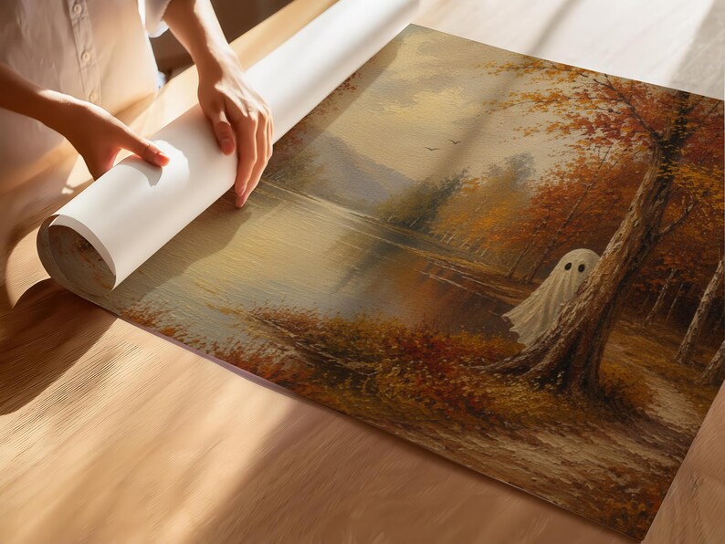 May include: A rectangular canvas print featuring an oil painting of an autumn landscape with a lake, orange and brown trees, and a small white ghost figure standing near a tree trunk. The artwork is being unrolled on a light wooden surface.