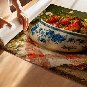 May include: A printed canvas wall art piece featuring a still life painting of a blue and white ceramic bowl filled with fresh red strawberries, resting on a white cloth with red checkered patterns, accented by small white daisy flowers.