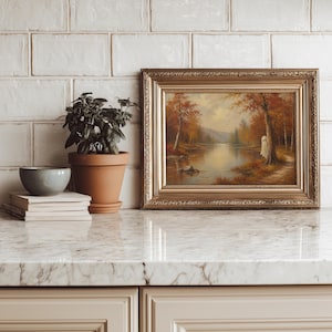 May include: A framed landscape painting featuring an autumn forest scene with a small white ghost figure standing by a tree near a calm lake. The artwork is enclosed in an ornate gold frame and sits next to a potted green plant and a stack of books.