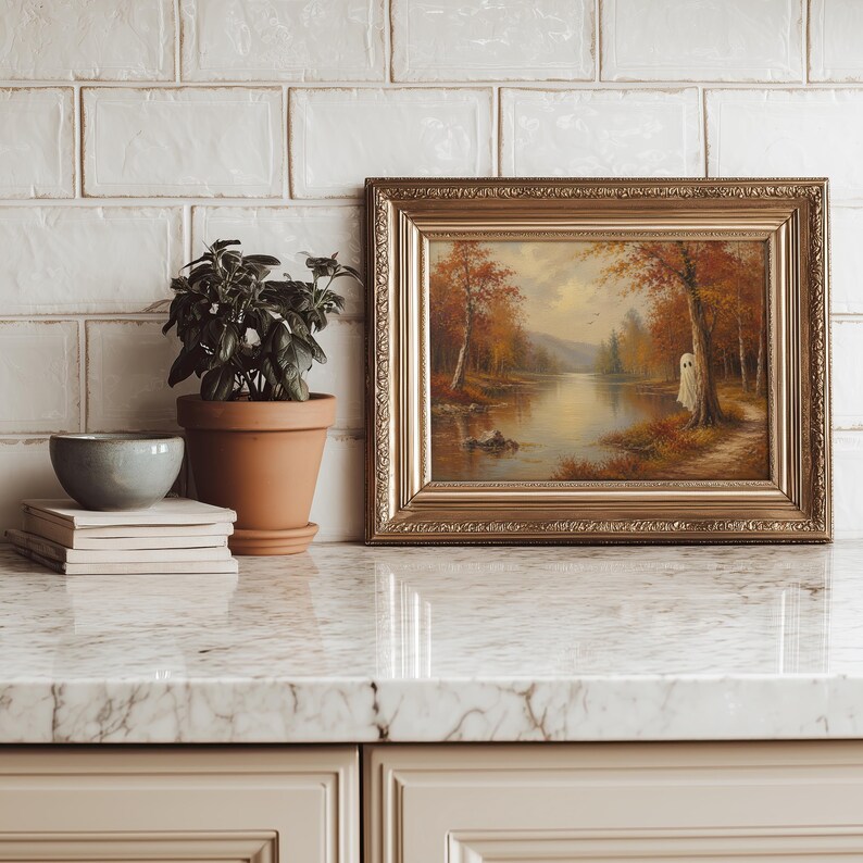 May include: A framed landscape painting featuring an autumn forest scene with a small white ghost figure standing by a tree near a calm lake. The artwork is enclosed in an ornate gold frame and sits next to a potted green plant and a stack of books.