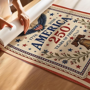 Puede incluir: Un tapiz beige con el texto "AMERICA 250 YEARS OF FREEDOM" y las fechas 1776 y 2026. El dise&ntilde;o incluye un &aacute;guila, estrellas y una campana. El tapiz tiene detalles en rojo, blanco y azul.