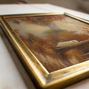 May include: A framed landscape painting featuring a white ghost figure resting on a fallen tree trunk within an autumn forest. The artwork displays warm orange and brown foliage, a calm body of water, and is encased in a metallic gold frame.