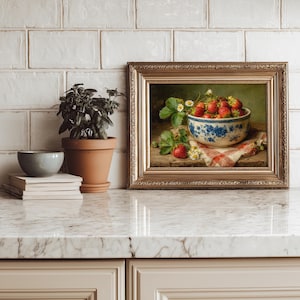 May include: A framed oil painting depicting a bowl of red strawberries with green leaves and small white flowers. The bowl features a blue floral pattern and sits on a red and white checkered cloth. The frame is ornate, metallic gold, and rectangular.
