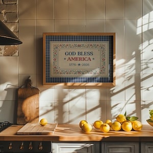 May include: A framed wall art piece with the text "GOD BLESS AMERICA" and the date "1776 - 2026". The artwork has a blue and white gingham border and floral accents. The image is in a kitchen setting with lemons.
