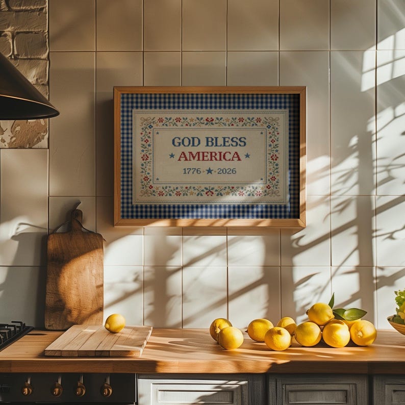 May include: A framed wall art piece with the text "GOD BLESS AMERICA" and the date "1776 - 2026". The artwork has a blue and white gingham border and floral accents. The image is in a kitchen setting with lemons.